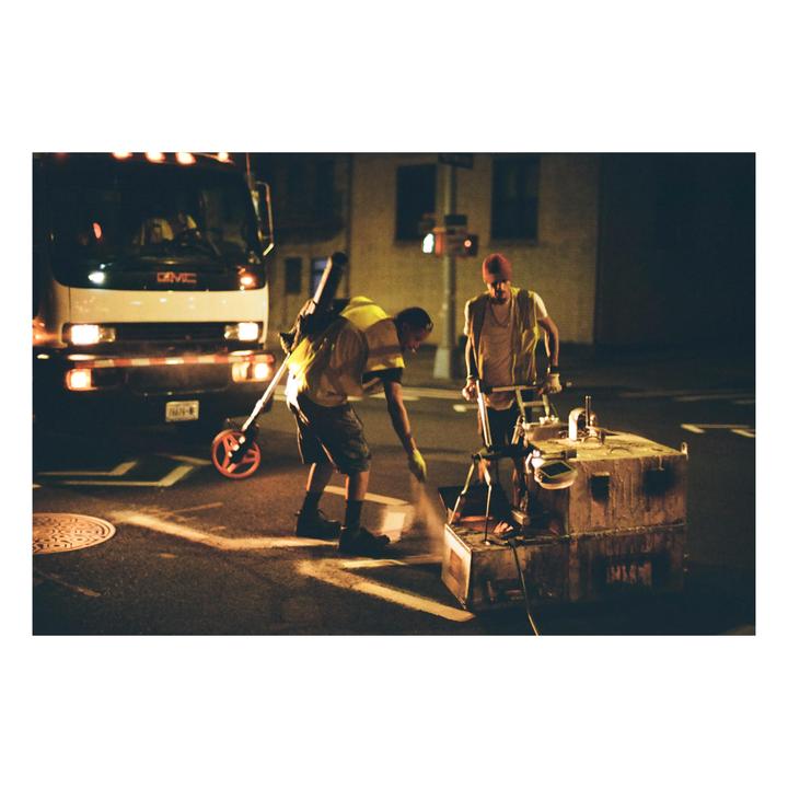 Two men in safety vests paint a white line on a street at night, with a truck parked behind them.