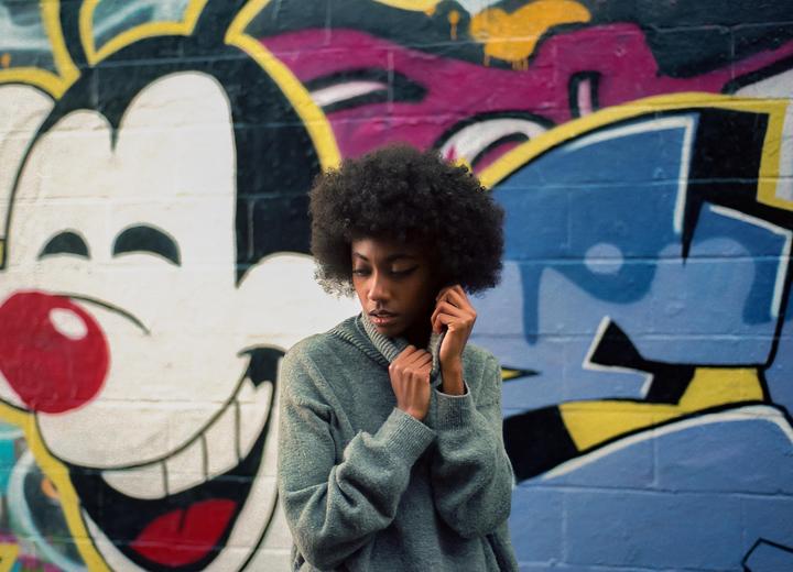 A woman with curly hair is looking down while holding her sweater in front of a colorful graffiti wall.