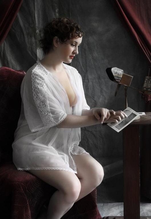 A woman in a sheer white robe sits on a red velvet chair, looking at a photograph.