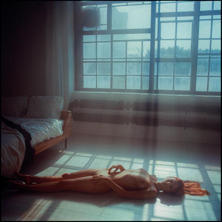A nude woman with red hair lies on the floor in a room with a large window and a bed.