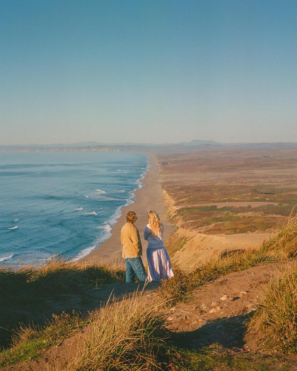 A couple stands on a grassy cliff overlooking a long sandy beach and the ocean.