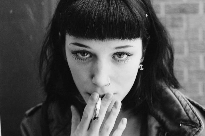 A woman with bangs and a nose ring holds a cigarette to her mouth.
