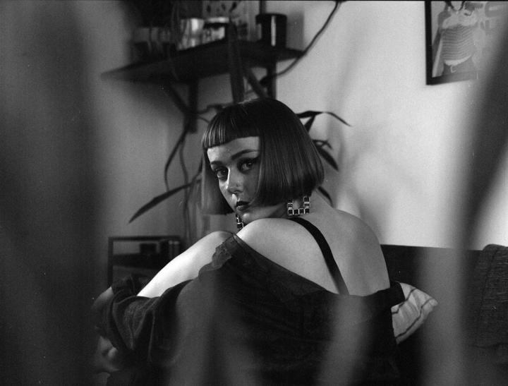 A woman with short hair and dark makeup looks over her shoulder at the viewer.