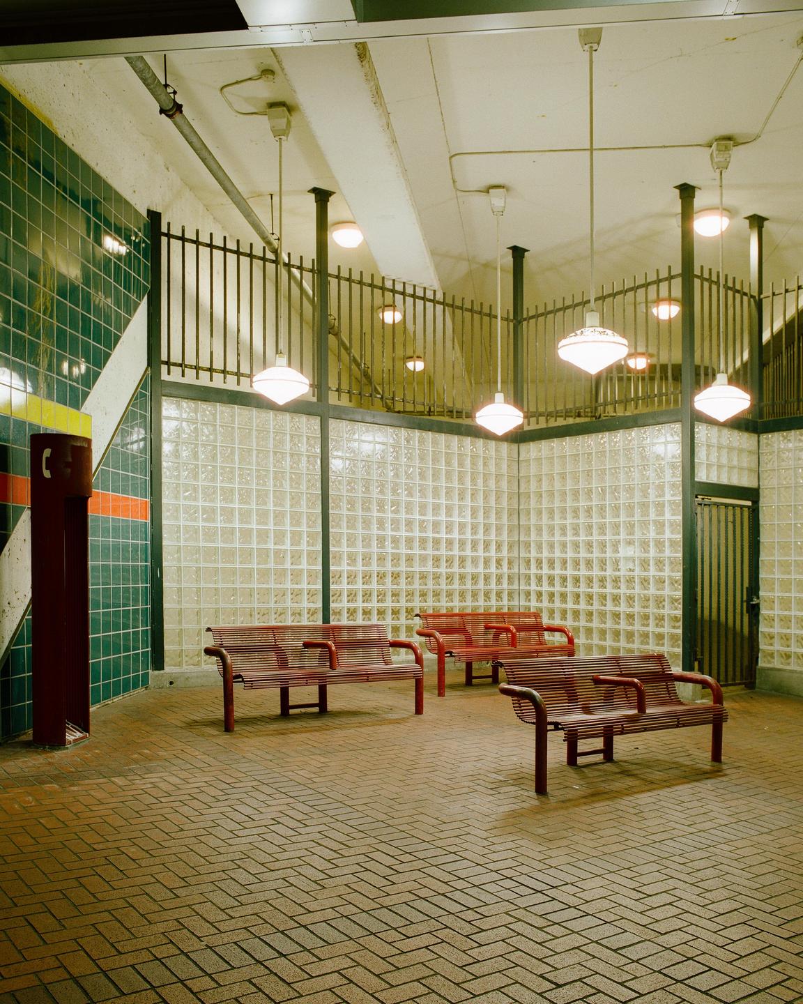 Three red benches sit on a brick floor in a waiting area with glass block walls and hanging lights.