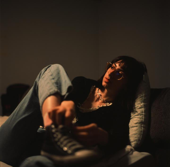 A woman wearing glasses and a necklace is lying on a couch, looking to the side, and holding a shoe.