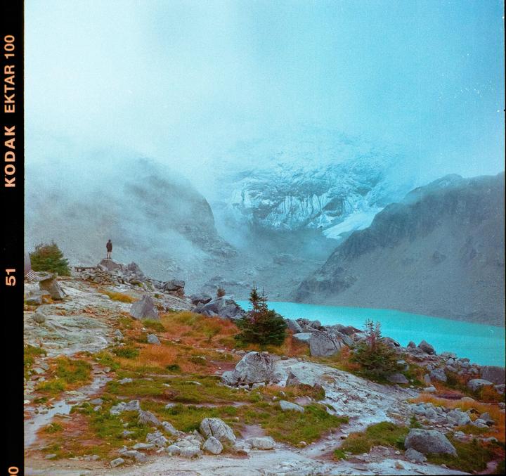 A person stands on a rocky outcrop overlooking a turquoise lake and misty mountains.