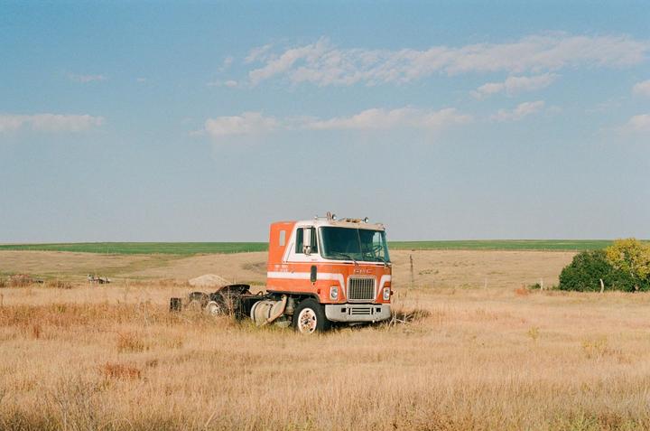 An orange and white semi-truck is parked in a field of dry grass under a blue sky with scattered clouds.