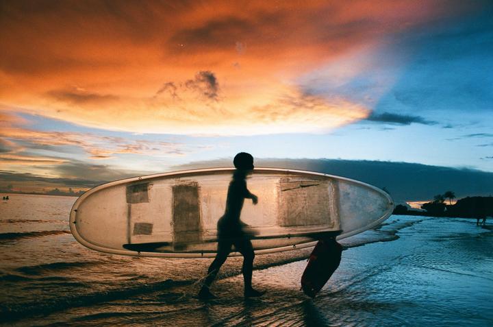 A person carries a boat and a paddle along the water's edge at sunset.