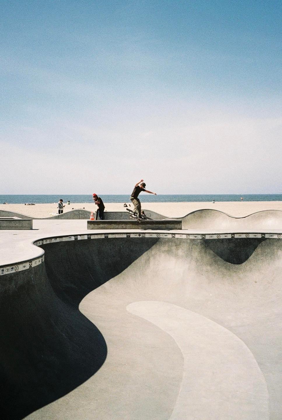 A skateboarder performs a trick in a concrete skatepark next to a beach.