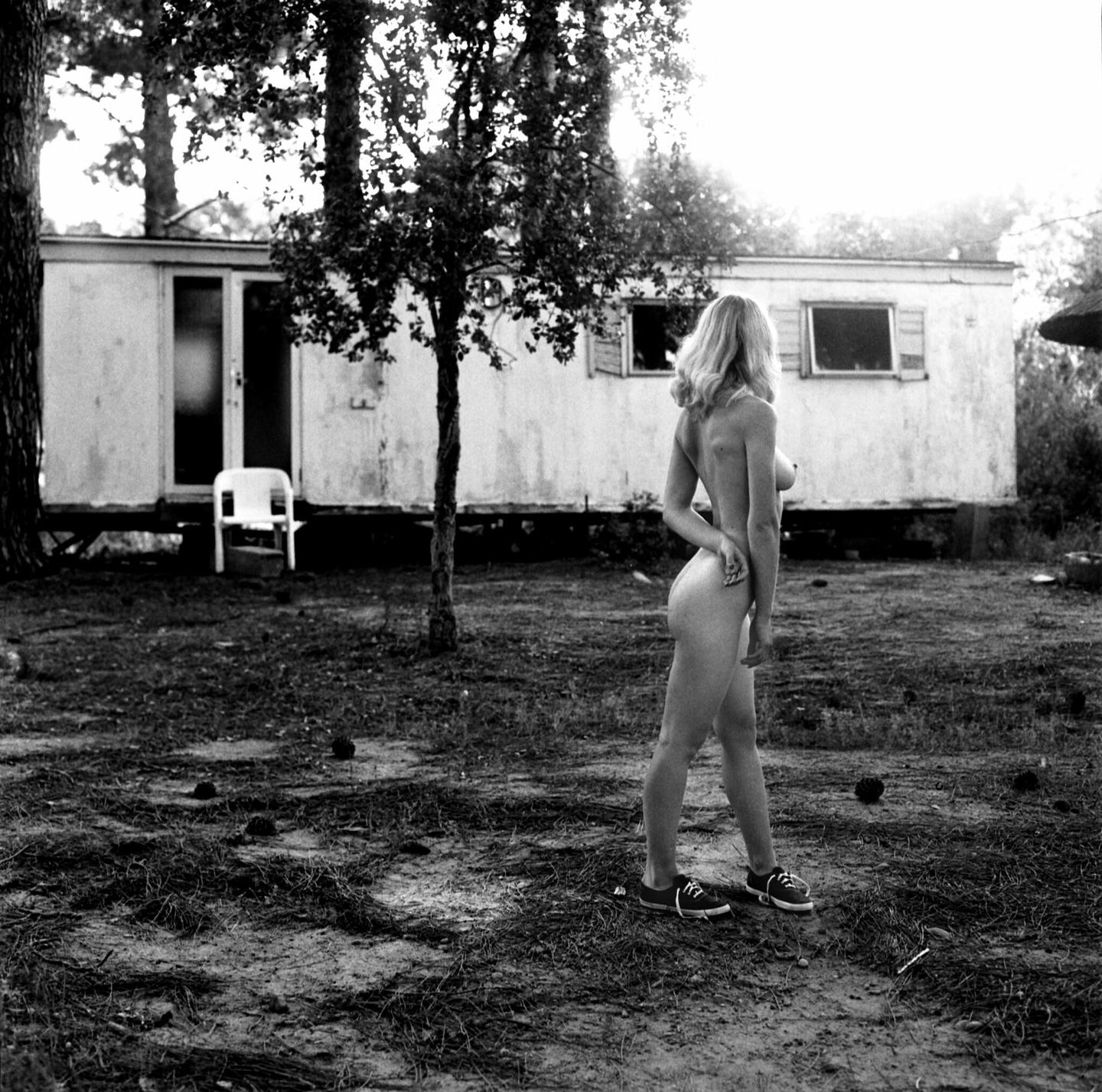 A nude woman stands in a yard with a trailer and trees in the background.