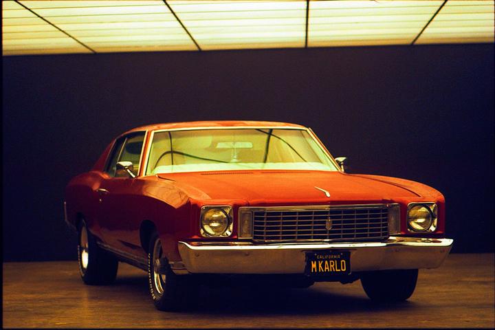 A red car with a California license plate that reads "M KARLO" is parked in a dark room with a grid of lights on the ceiling.