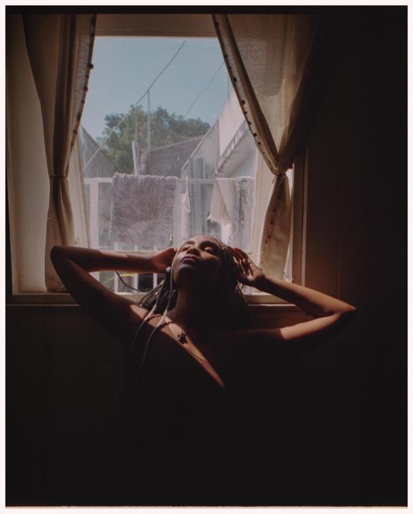 A woman with braided hair and a necklace is bathed in sunlight from a window, with her arms raised behind her head.