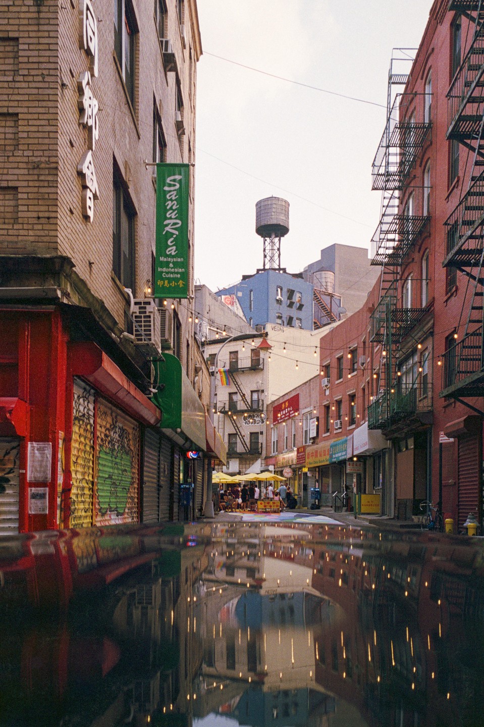 A street in New York City is reflected in a puddle, with buildings and a water tower visible.
