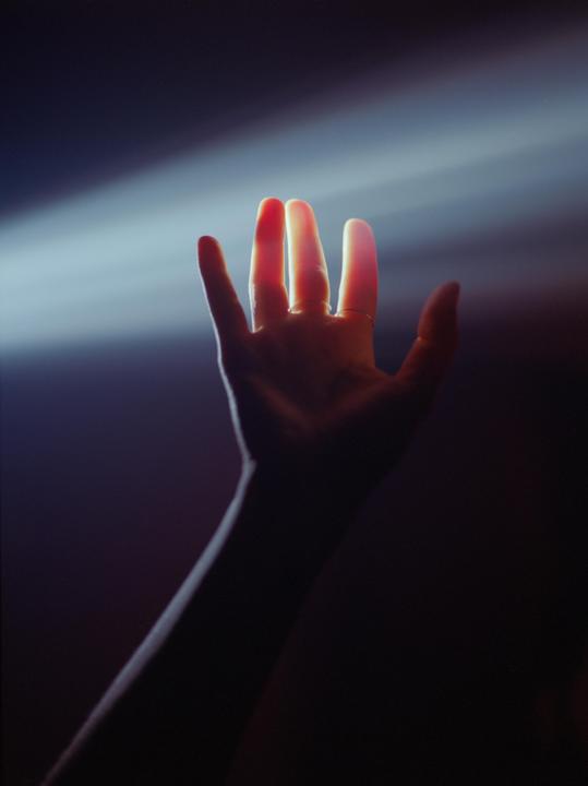 A hand with rings is backlit by a bright light beam against a dark background.