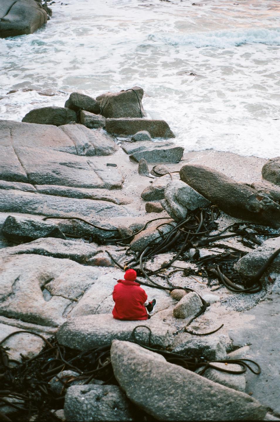 A person in a red jacket sits on a rock at the edge of a beach with waves crashing nearby.