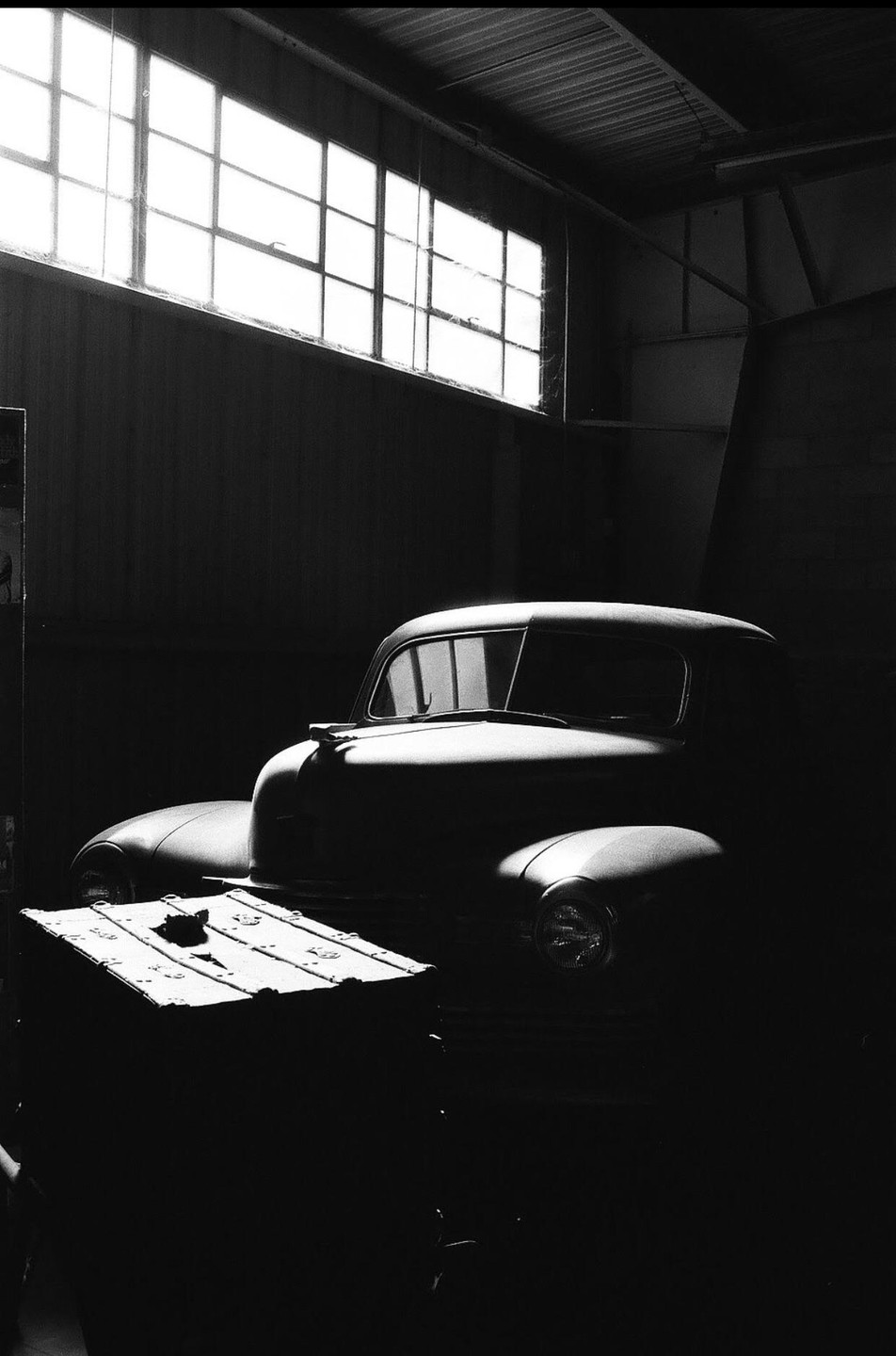 A vintage car is parked in a dark garage with a bright window above it.