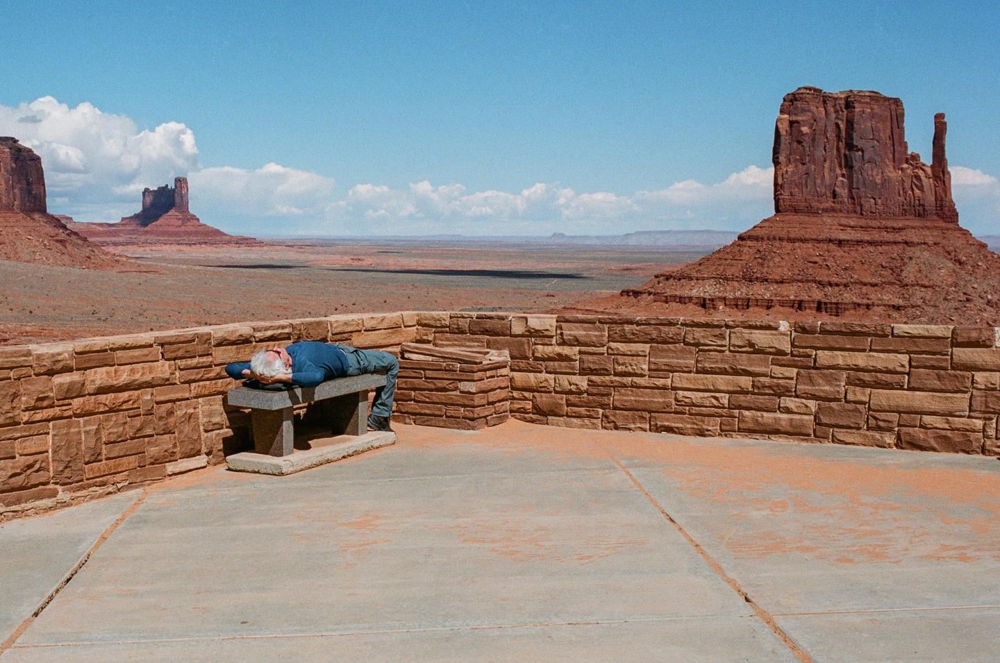 A man sleeps on a stone bench overlooking a desert landscape with large rock formations under a blue sky.
