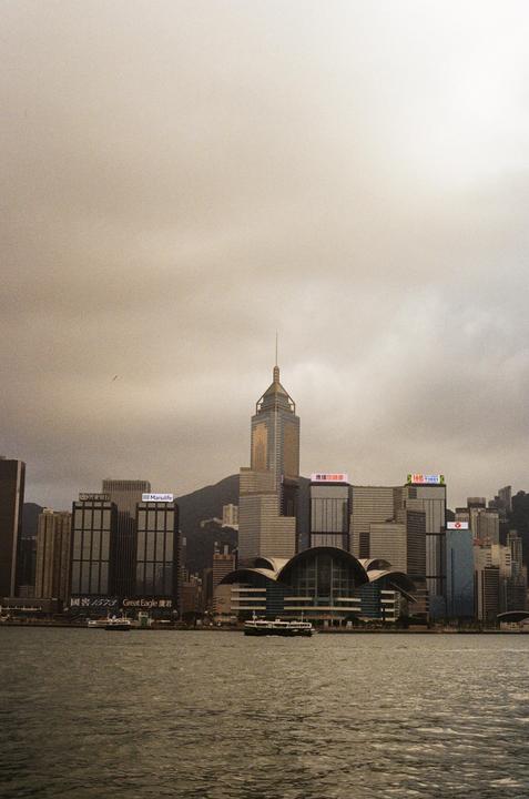 A cityscape with tall buildings and a body of water in the foreground is visible under a cloudy sky.