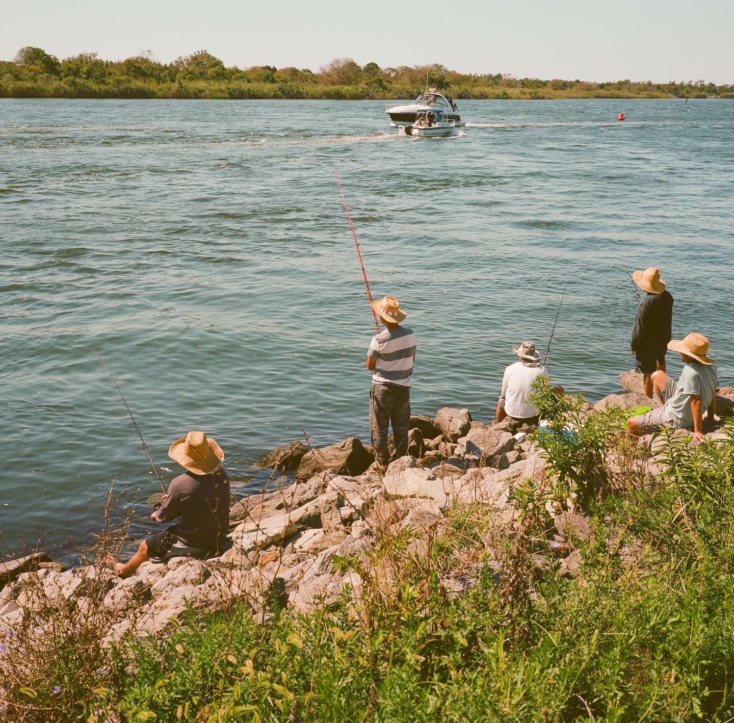 Four men wearing straw hats are fishing from a rocky shore as a boat passes by on the water.