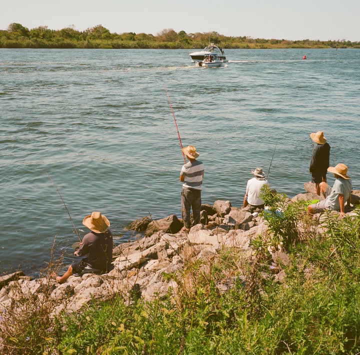 Four men wearing straw hats are fishing from a rocky shore as a boat passes by on the water.