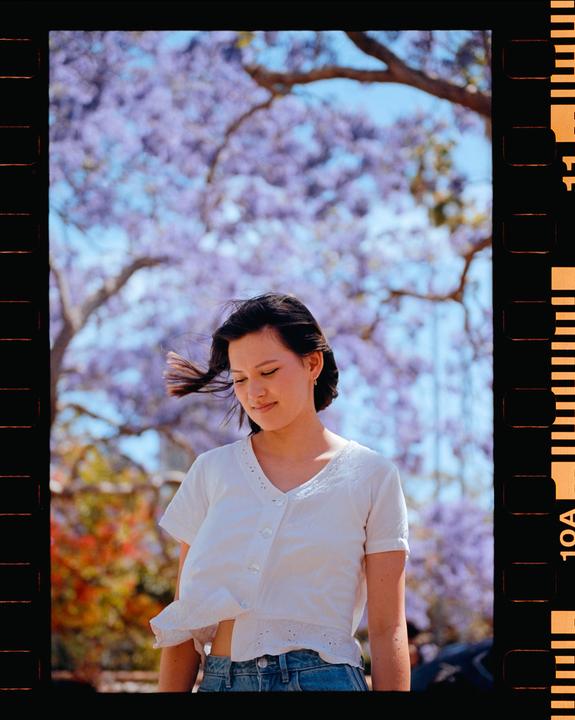 A woman stands outdoors with purple jacaranda blossoms in the background.