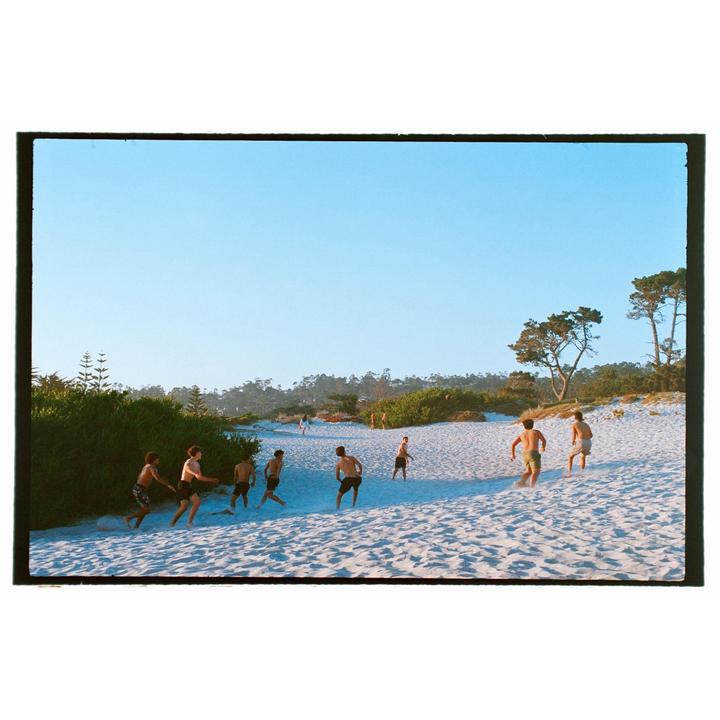 A group of young men play on a sandy beach with trees and bushes in the background under a clear sky.