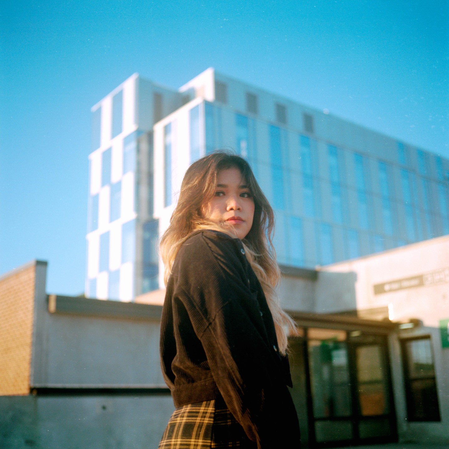 A woman with long hair looks over her shoulder at the viewer, with a modern building in the background.