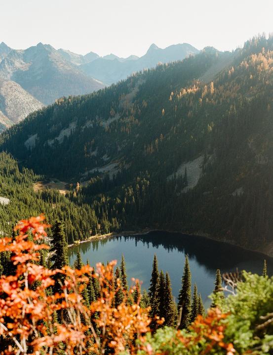 A mountain lake is surrounded by pine trees and autumn foliage under a clear sky.