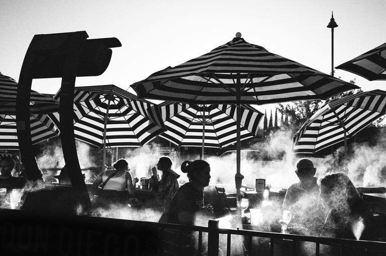 People sit at tables under striped umbrellas on a hazy summer evening.
