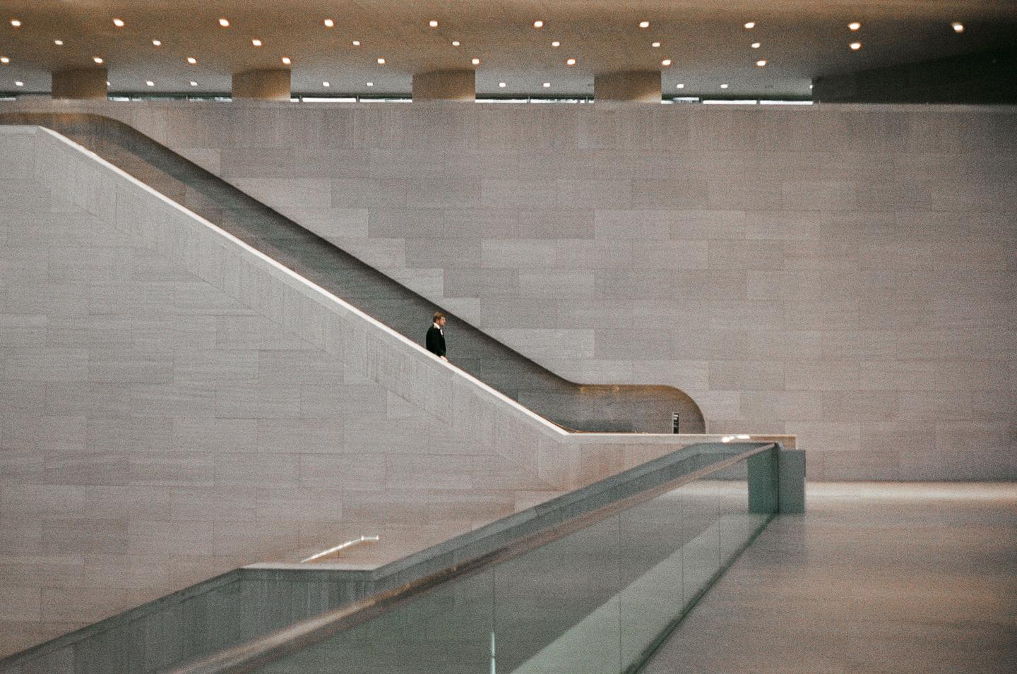 A man stands on an escalator in a modern building with stone walls and a patterned ceiling.