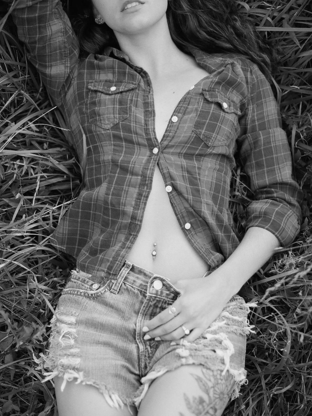 A woman wearing an unbuttoned plaid shirt and ripped denim shorts lies in tall grass.
