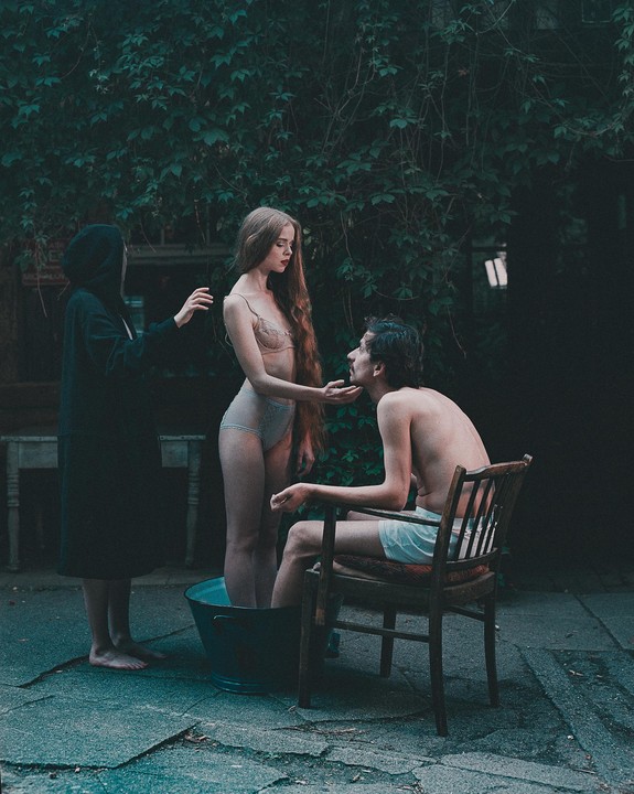 A woman in lingerie stands in a tub while holding the face of a man sitting in a chair, as another person in a hooded robe stands nearby.