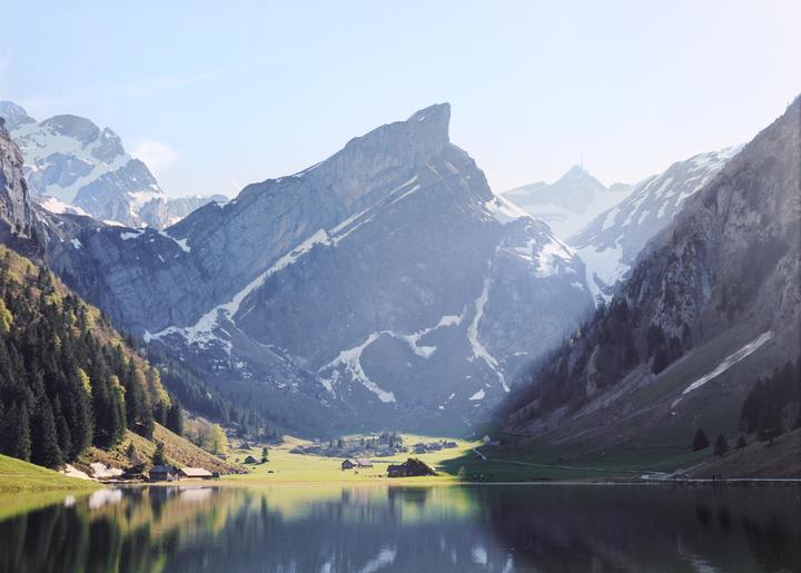 A lake reflects a valley with houses and snow-capped mountains under a bright sky.