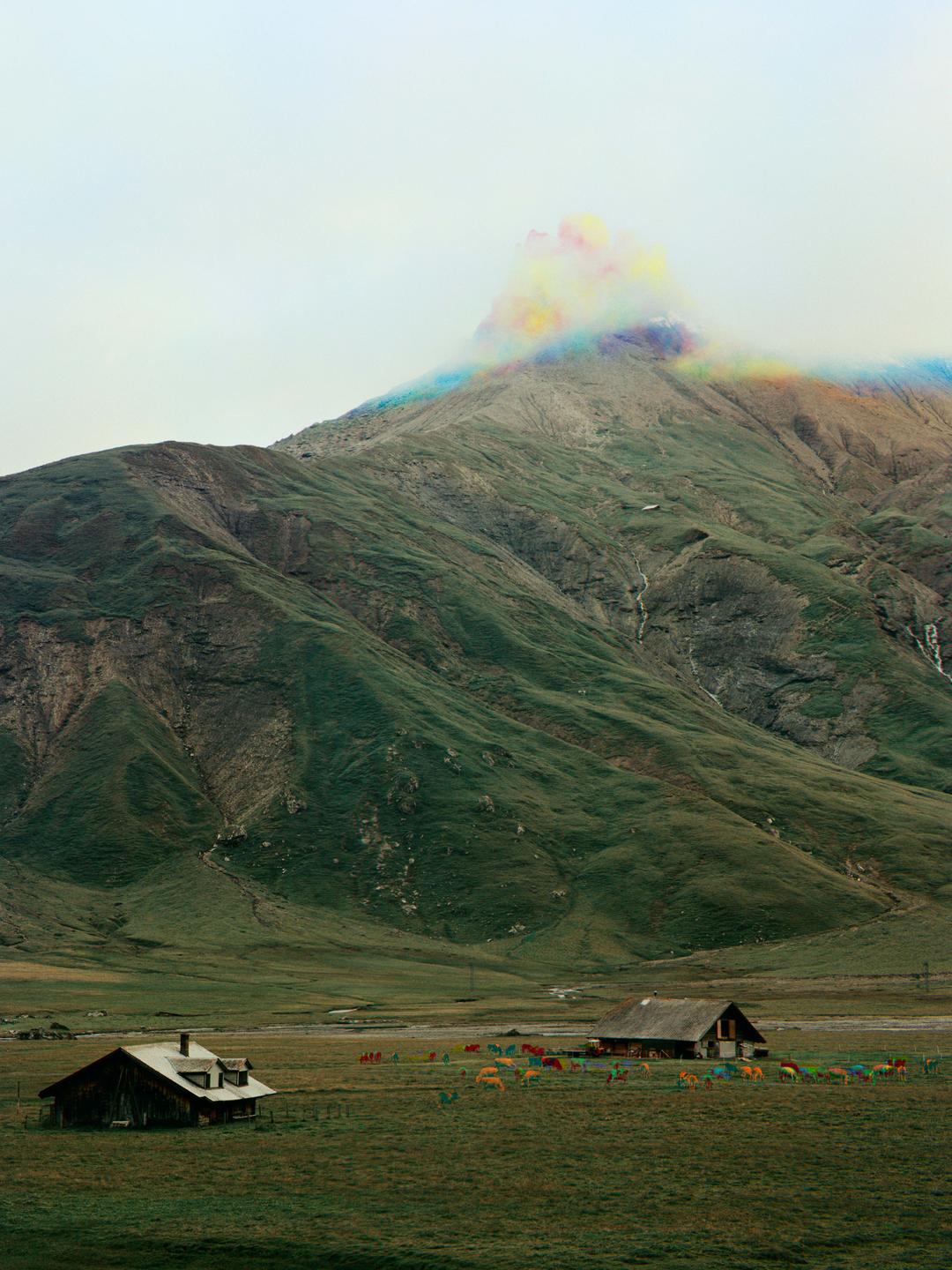 A colorful cloud hovers over a mountain with two houses and a herd of cattle in the foreground.