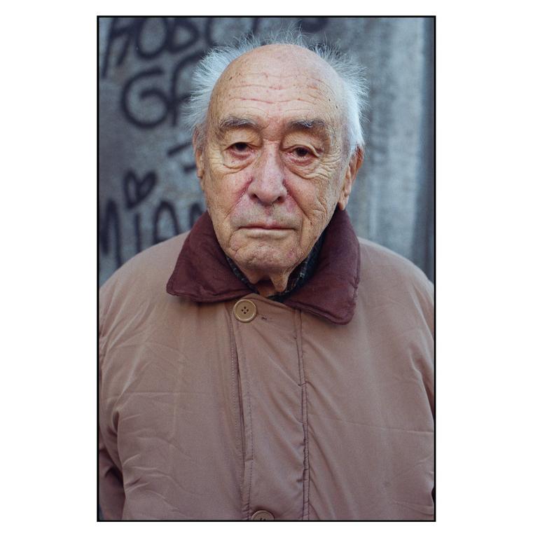 A man with white hair and a tan jacket stands in front of a wall with graffiti.