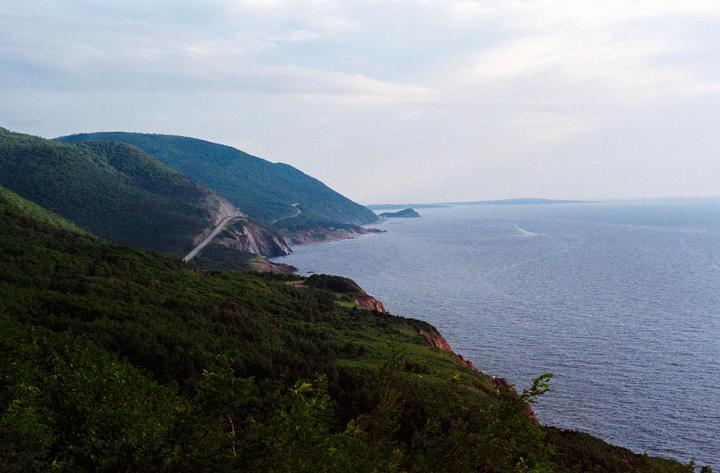 A winding road follows the coastline along a forested mountain range next to a large body of water.