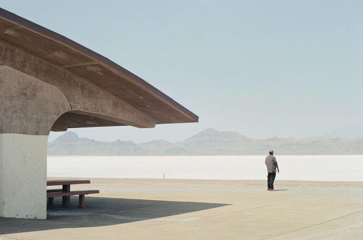 A man stands on a concrete slab looking out at a vast, flat, white landscape with mountains in the distance, next to a concrete shelter with a picnic table.