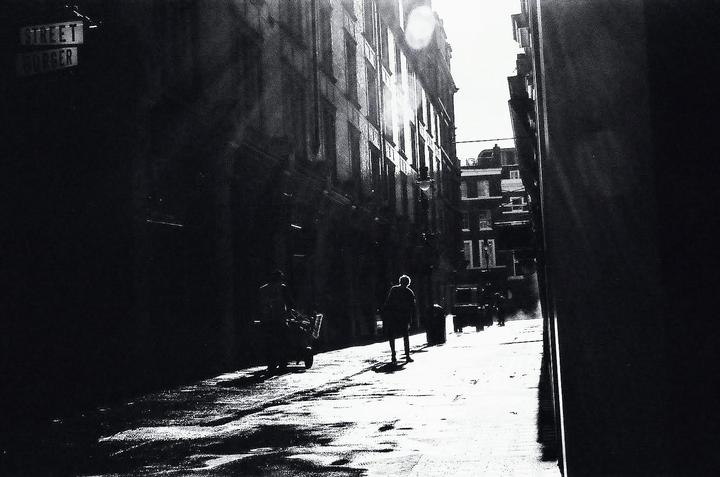A person walks down a sunlit street between tall buildings, with another person pushing a cart in the foreground.