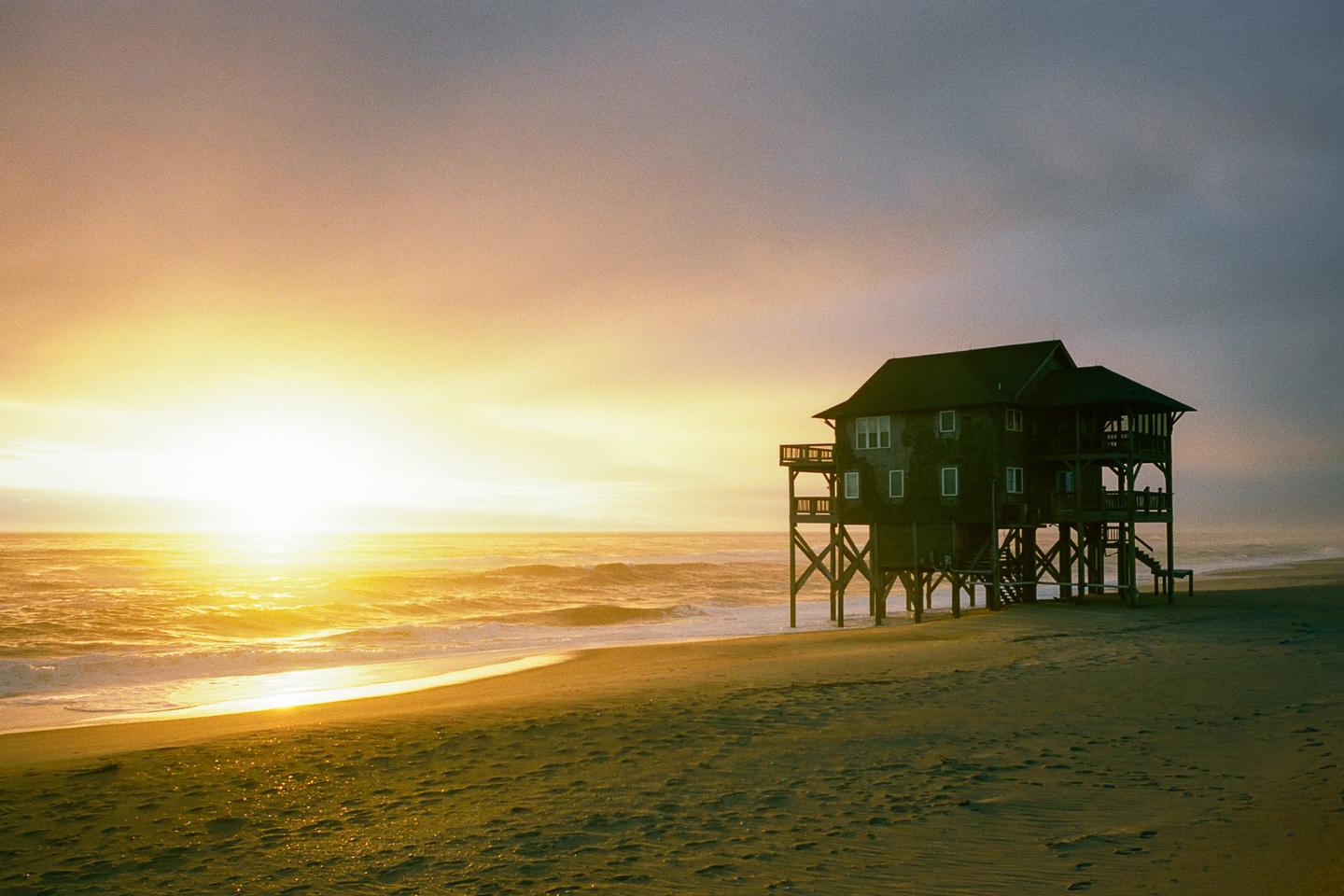 A house on stilts stands on a beach with waves crashing as the sun sets over the ocean.