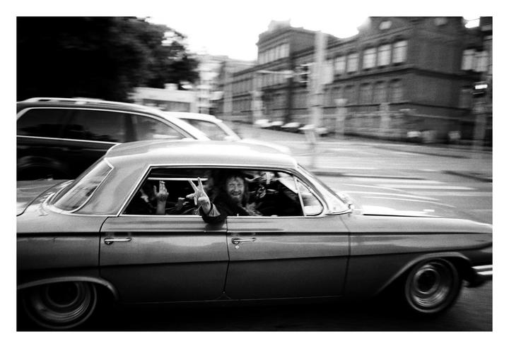 A man with a beard smiles and makes a peace sign from the window of a moving car on a city street.