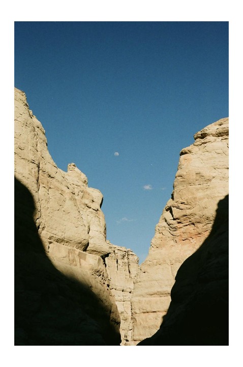 A moon is visible in the clear blue sky above a canyon with steep rock walls.