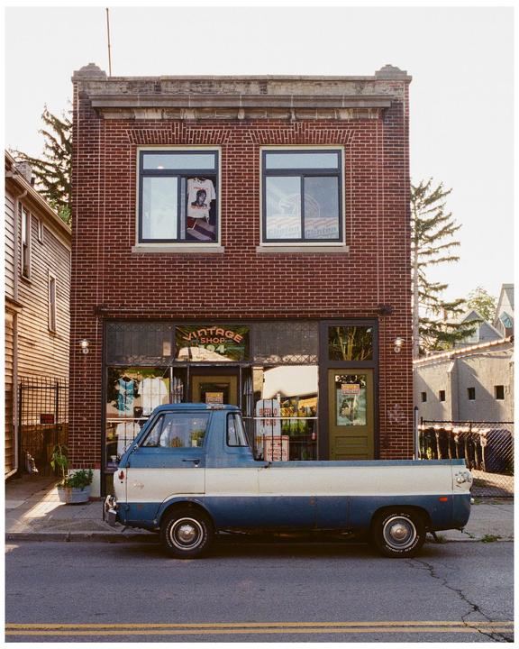 A blue and white vintage pickup truck is parked on a street in front of a brick building with a vintage shop.