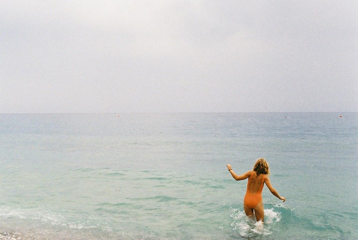 A woman in an orange swimsuit walks into the ocean from a rocky beach.