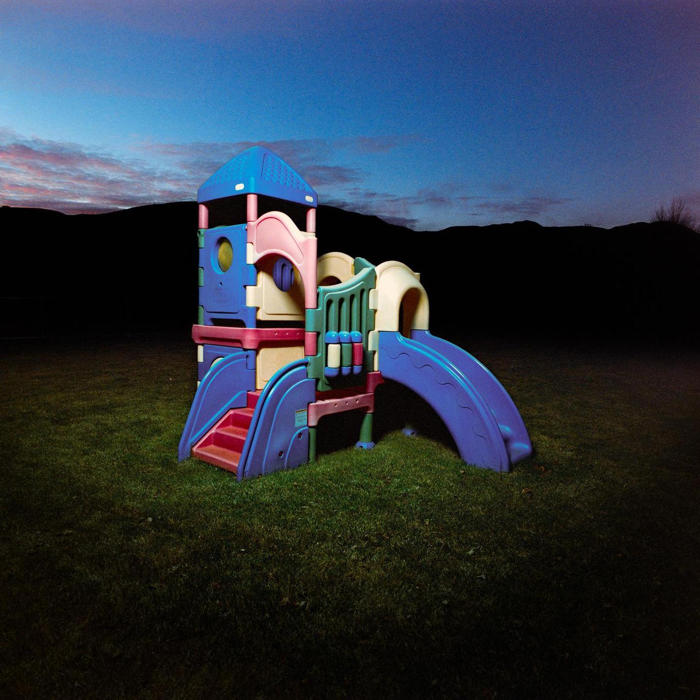 A colorful plastic playground structure sits on a grassy lawn under a twilight sky with a dark mountain range in the background.