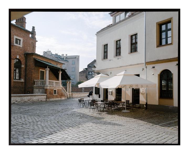 A cobblestone street is lined with buildings and an outdoor cafe with white umbrellas.