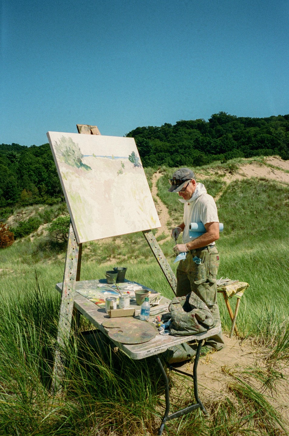 A man paints a landscape on an easel set up in a grassy, sandy area with trees in the background.
