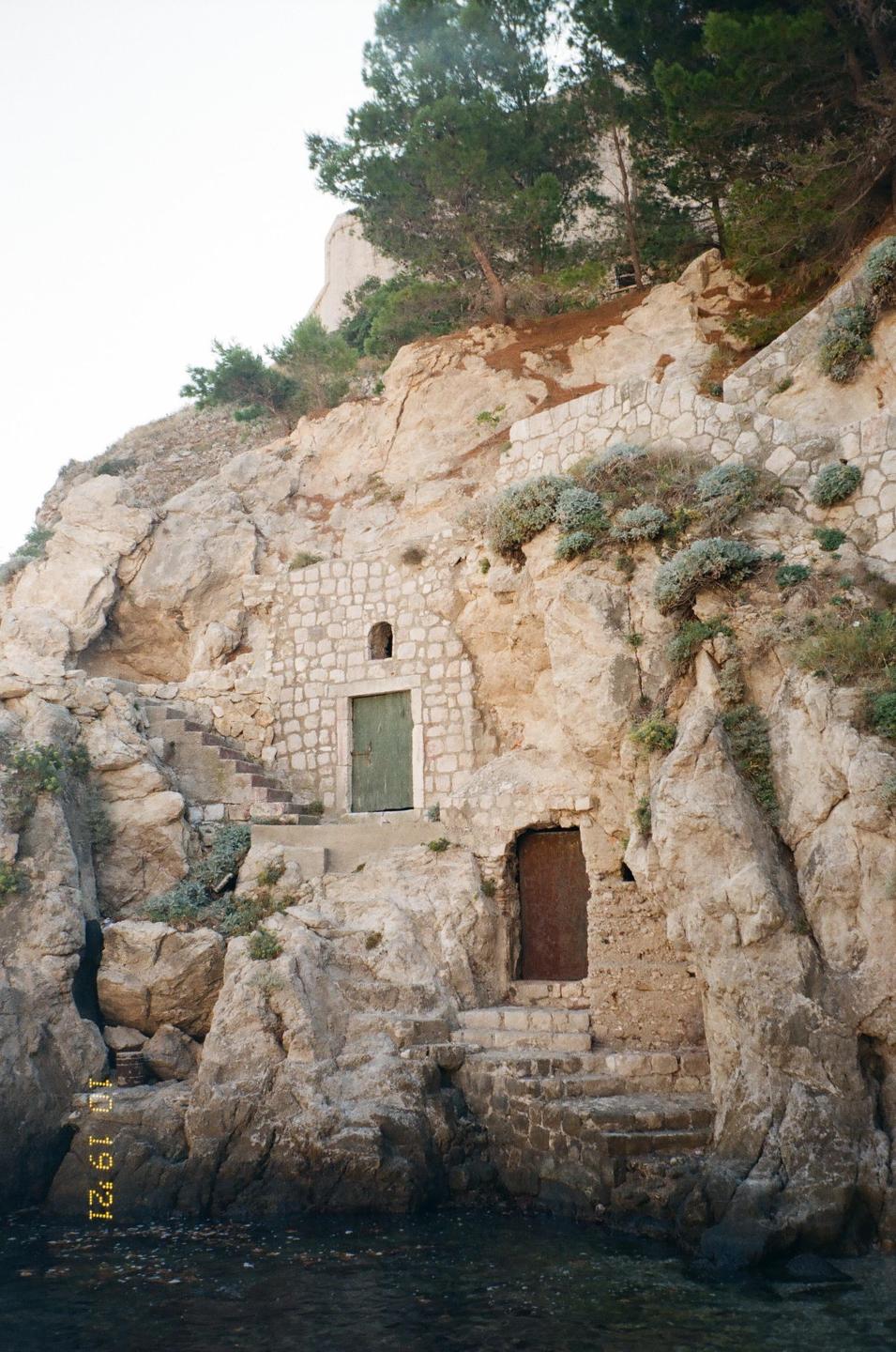 Two doors are built into a rocky cliffside above the water, with a stone staircase leading up to the top of the cliff.