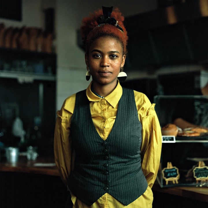 A woman with red hair and a yellow shirt with a pinstriped vest stands in what appears to be a cafe.