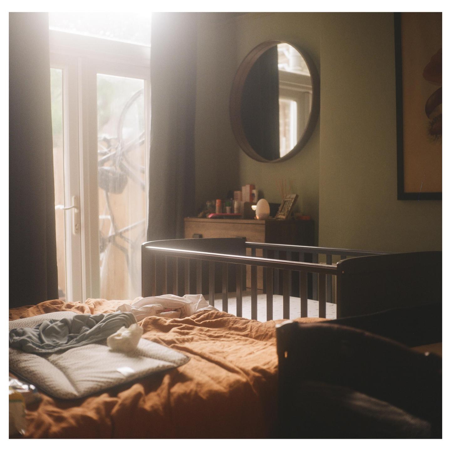 A baby's crib stands in a sunlit room with a bed in the foreground.