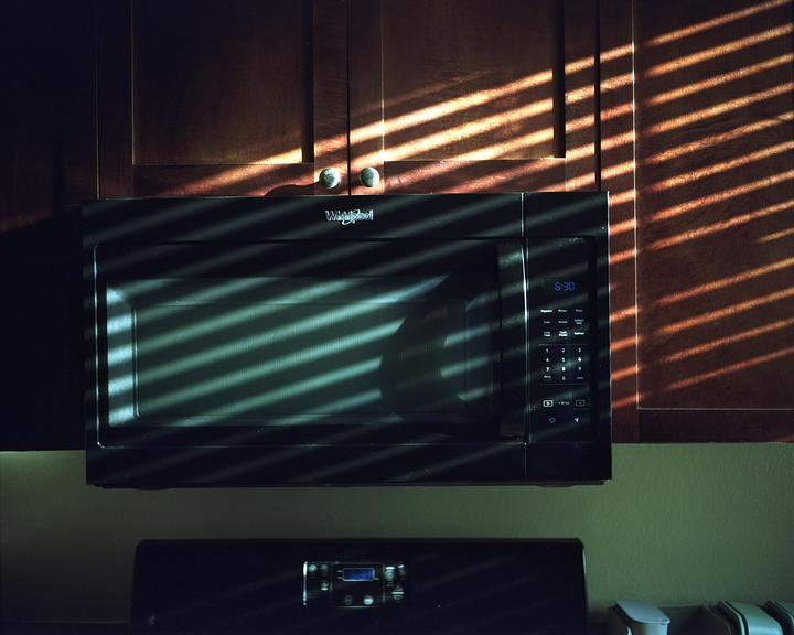 Sunlight streams through blinds, casting striped shadows across a microwave and kitchen cabinets.